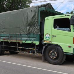 A light green Hino commercial truck with a dark green tarp-covered cargo bed parked along a roadside.
